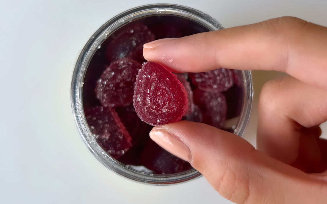 Top view of a red sugar-coated gummy held between fingers above an open jar on a white background
