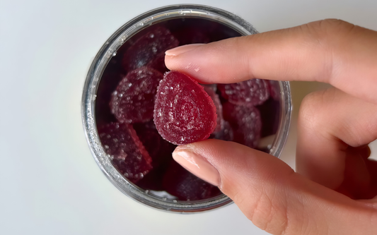 Top view of a red sugar-coated gummy held between fingers above an open jar on a white background