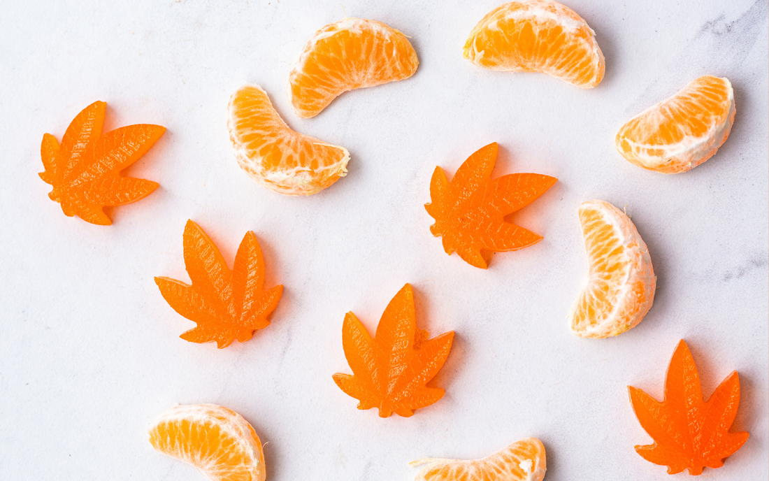 Orange cannabis leaf gummy candies arranged with peeled mandarin segments on a white marble background