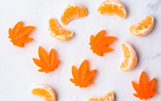 Orange cannabis leaf gummy candies arranged with peeled mandarin segments on a white marble background