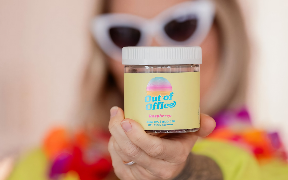 Close-up front view of Out of Office raspberry edible jar showing yellow label and flavor, held by hand with blurred person in sunglasses behind