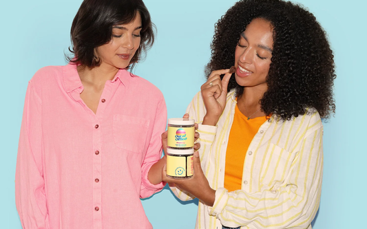 Two women holding jars of “Out of Office” gummies as one brings a gummy to her mouth against a light blue background