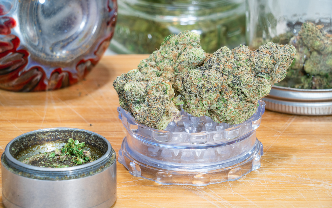 Close-up of cannabis buds on a clear grinder, showing dense frosty nugs and orange pistils with a wooden board and jar behind