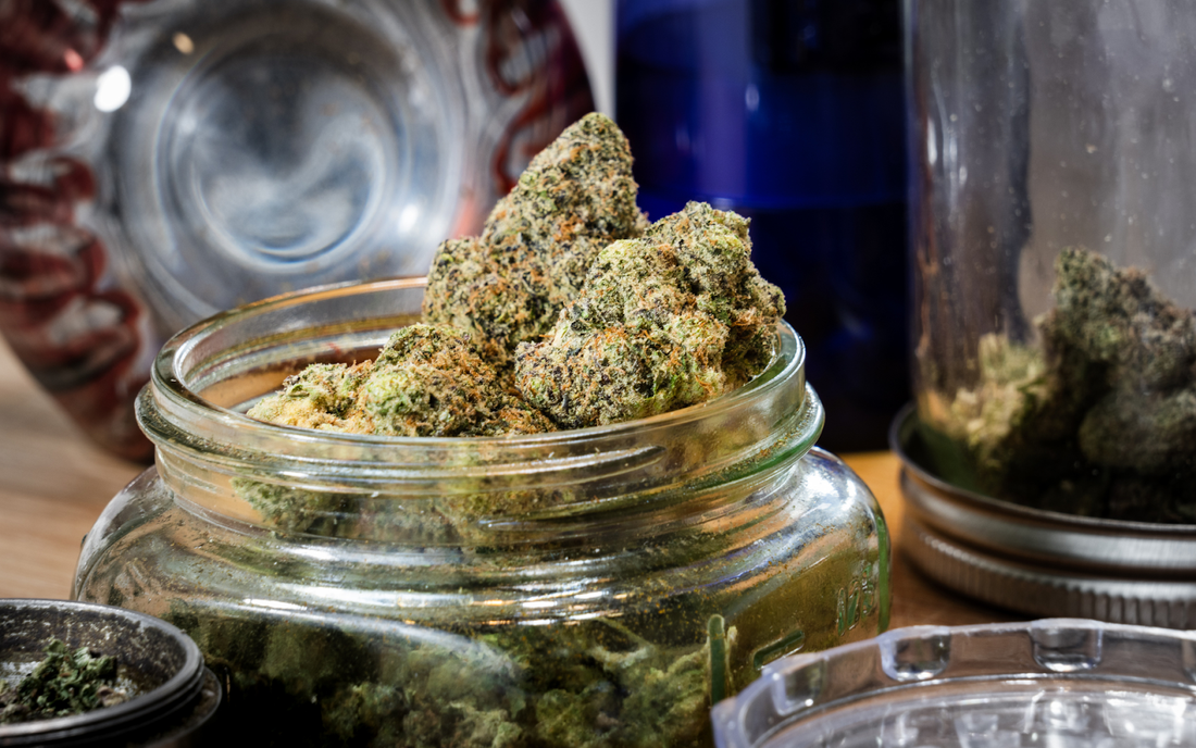Close-up of cannabis buds in a glass jar on a wooden table with a grinder and other jars in the background