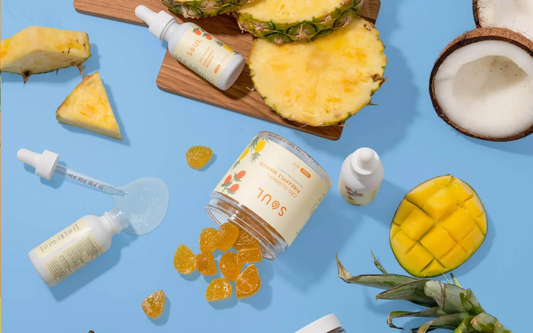 Flat lay of Soul CBD pineapple-mango gummies jar and tincture bottles with spilled gummies and fresh pineapple, mango, and coconut on a blue background