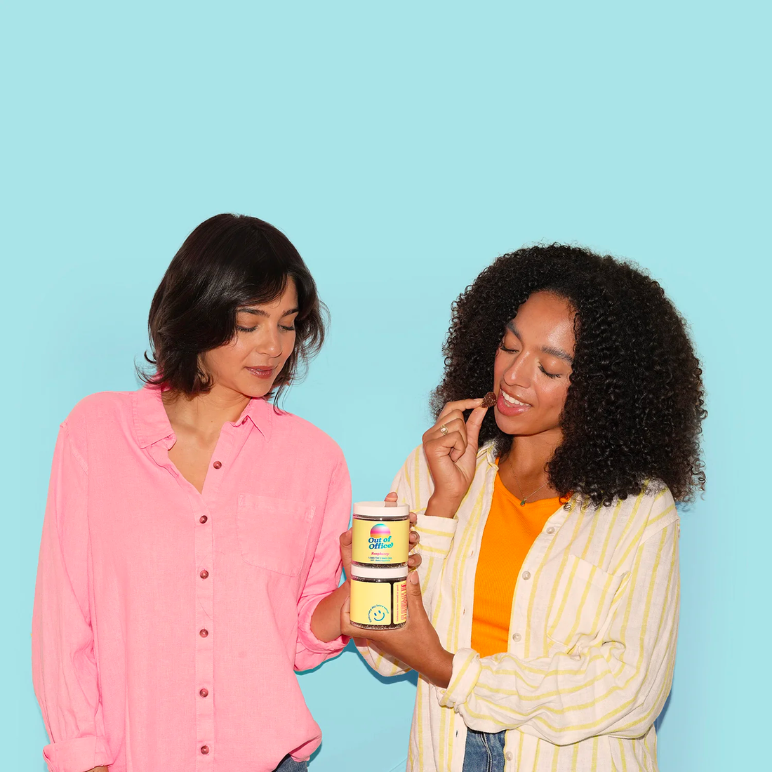 Two women sample a gummy while holding stacked product jars against a light blue background