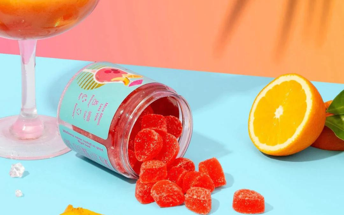 Angled view of a tipped jar of red sugar-coated CBD gummies spilling onto a blue surface, with a sliced orange and peach-colored background