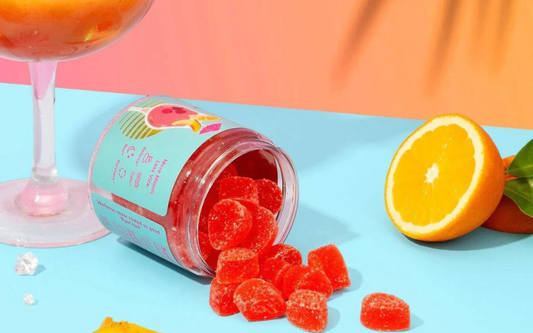 Angled view of a tipped jar of red sugar-coated CBD gummies spilling onto a blue surface, with a sliced orange and peach-colored background