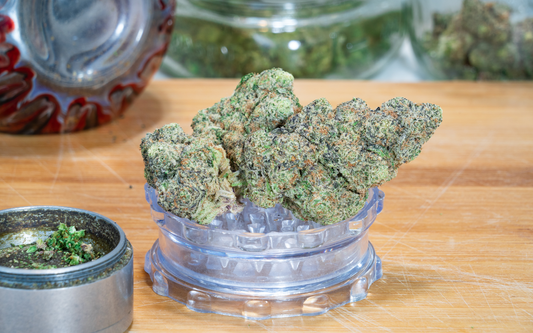 Close-up of cannabis buds resting on a clear grinder lid on a wooden surface with a metal grinder and glass jars blurred in the background