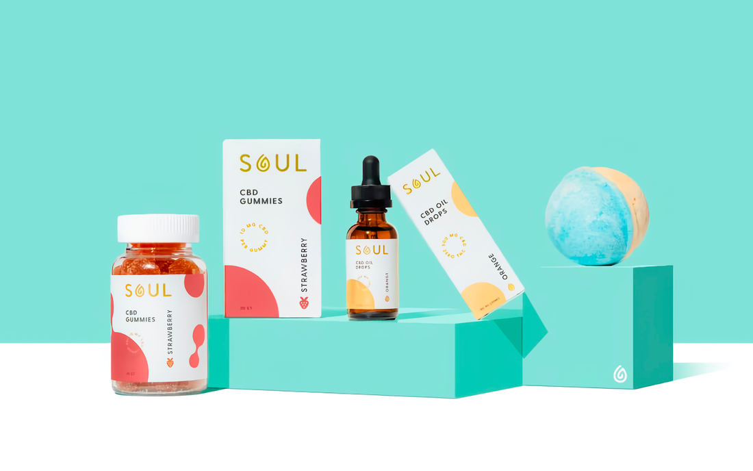 Front view of Soul CBD product lineup showing a jar of strawberry CBD gummies, boxed gummies, a CBD oil dropper bottle, and a blue bath bomb on teal pedestals