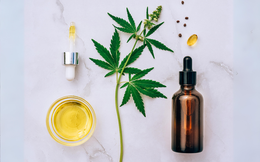 Cannabis leaf with amber dropper bottle, glass bowl of yellow oil, and a softgel capsule arranged on a white marble surface