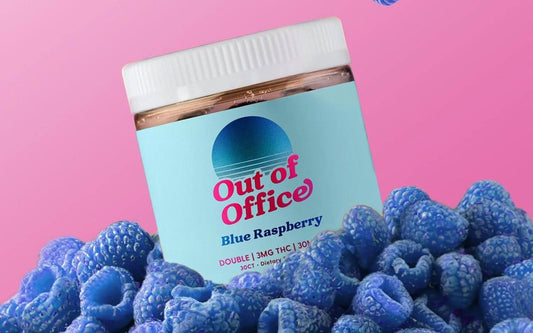 Tilted jar of Out of Office Blue Raspberry gummies showing a pastel blue label and white lid, surrounded by blue raspberries on a pink background