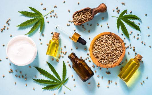 Flat lay of hemp products: hemp leaves, a wooden bowl of hemp seeds, a jar of cream, and small oil bottles with dropper on a blue background