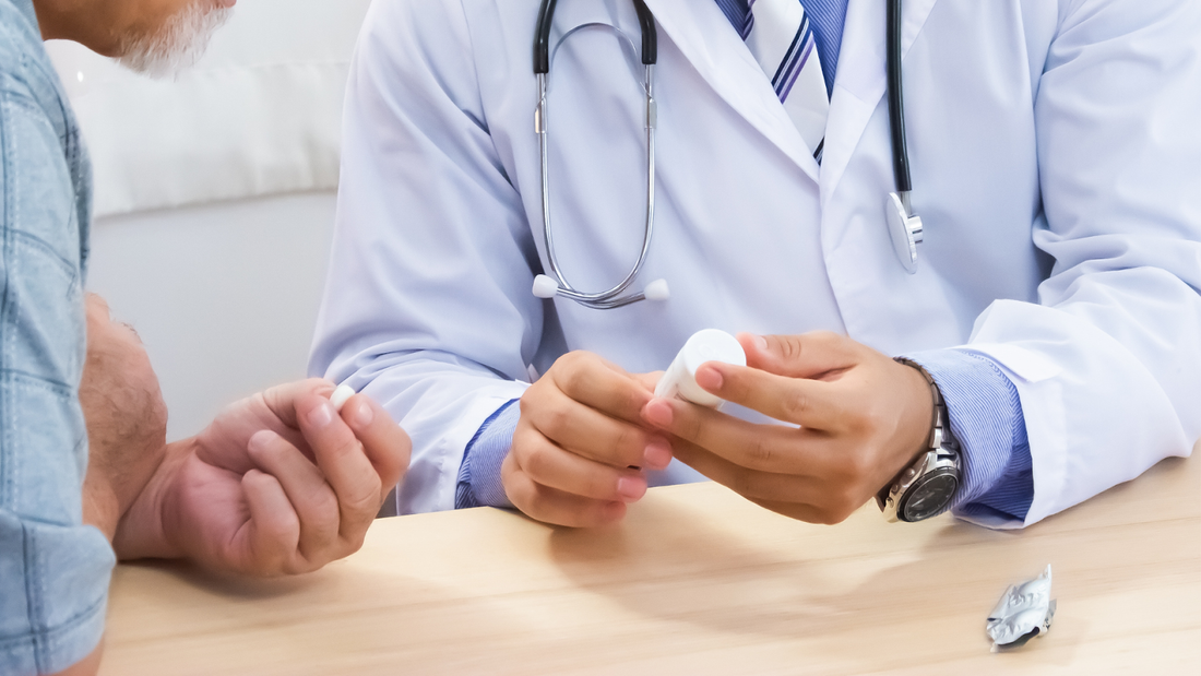 Doctor in white coat with stethoscope explaining a pill bottle to an older patient at a clinic table