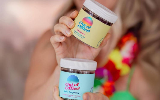 Close up of two Out of Office gummy jars showing yellow Raspberry and blue Blue Raspberry labels and white caps against a blurred colorful background