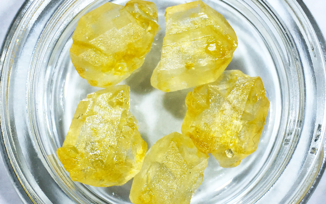Close-up of THC-JD crystal chunks in a glass jar showcasing translucent yellow-amber color and rough faceted texture on white background