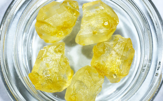 Close-up of THC-JD crystal chunks in a glass jar showcasing translucent yellow-amber color and rough faceted texture on white background