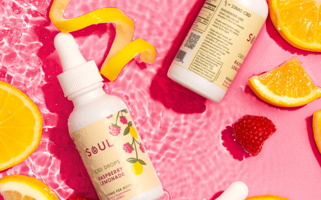 Angled bottle of Soul Raspberry Lemonade CBD drops showing dropper top and label with sliced oranges and raspberries on pink background