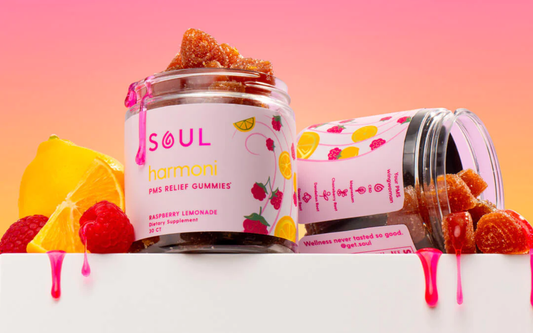 Angled front view of Soul Harmoni Raspberry Lemonade PMS Relief Gummies jars, one tipped spilling sugar-coated gummies with lemon and raspberries on a pink-orange gradient background