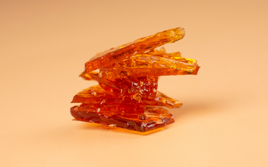 Close-up side view of stacked amber cannabis concentrate shards (shatter) showcasing glossy translucent texture on peach background