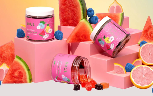 Three-quarter view of Soul CBD Gummies jars, one tipped spilling sour gummies on pink stepped display with watermelon and grapefruit props