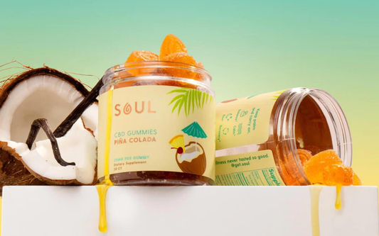 Front view of Soul Pina Colada CBD gummies jar showing orange sugar-coated gummies spilling beside halved coconut on a tropical gradient background