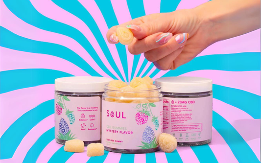 Front view of Soul CBD Gummies jar showing Mystery Flavor label and sugar-coated yellow gummies, hand holding one against pink and teal spiral background