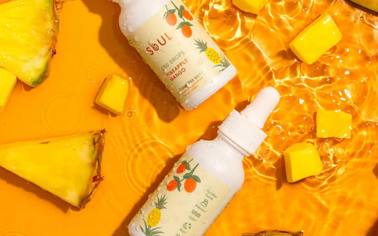 Top-down view of two Soul Pineapple Mango CBD oil dropper bottles with condensation, pineapple pieces on bright orange background