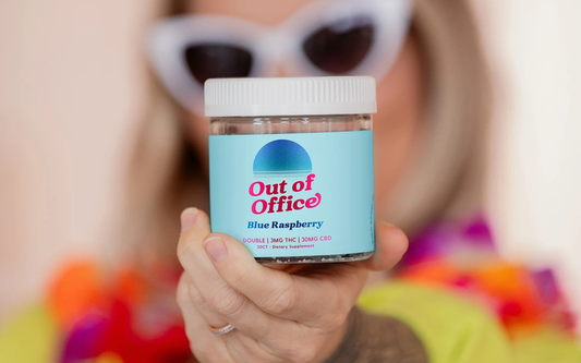 Close-up front view of Out of Office Blue Raspberry gummy jar showing blue label reading Out of Office and 3mg THC and 30mg CBD, held by a hand with a blurred person in the background