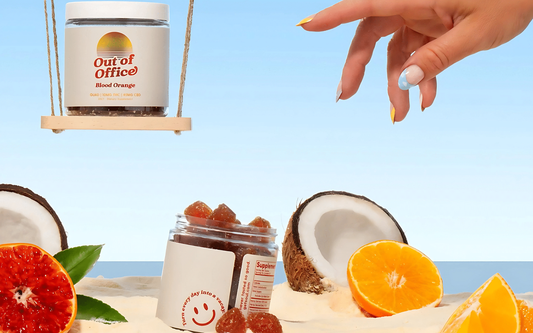 Out of Office Blood Orange THC gummies jar on a small swing and an open jar of gummies on sand with halved coconut and orange slices, hand picking a gummy