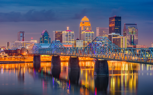 Downtown Louisville skyline at dusk with an illuminated truss bridge reflecting city lights in the river