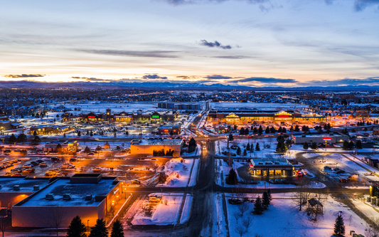 Aerial view of a Montana town at dusk with snow-covered streets, glowing shopping centers, and distant mountains