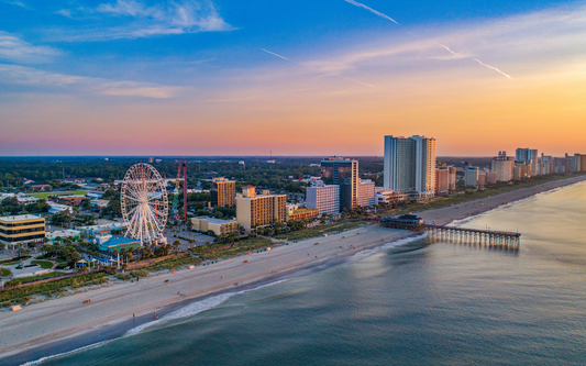 Aerial view of a coastal beach at sunset with a Ferris wheel, high-rise hotels, and a pier
