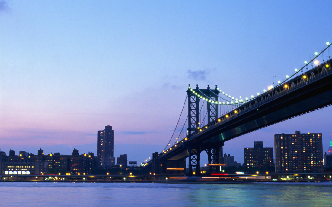 Suspension bridge lit at dusk spanning a river with city skyline and apartment buildings under a purple sky
