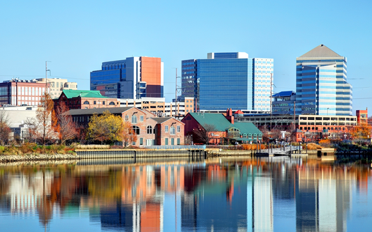 City skyline reflected in a calm river, showing glass office towers and brick waterfront buildings under a clear blue sky