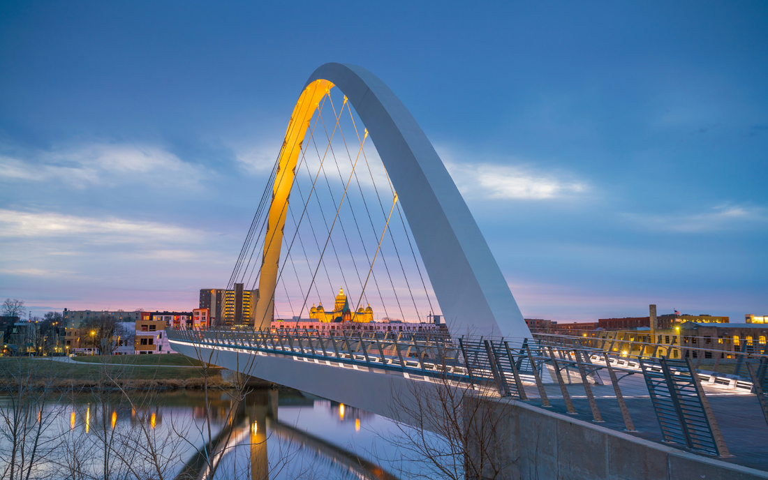 Pedestrian arch bridge at dusk over a river, city skyline with an illuminated domed building in the background
