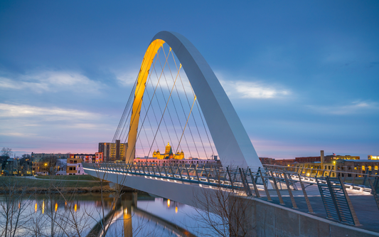 Pedestrian arch bridge at dusk over a river, city skyline with an illuminated domed building in the background