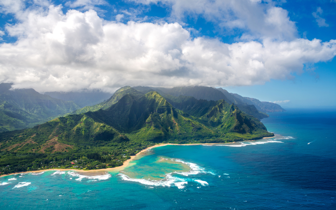 Aerial view of a Hawaiian coastline showing turquoise water, sandy beaches, and steep green mountains under low clouds