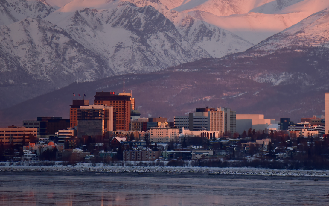 Anchorage skyline at sunset with snow-covered Chugach Mountains and a frozen inlet in the foreground