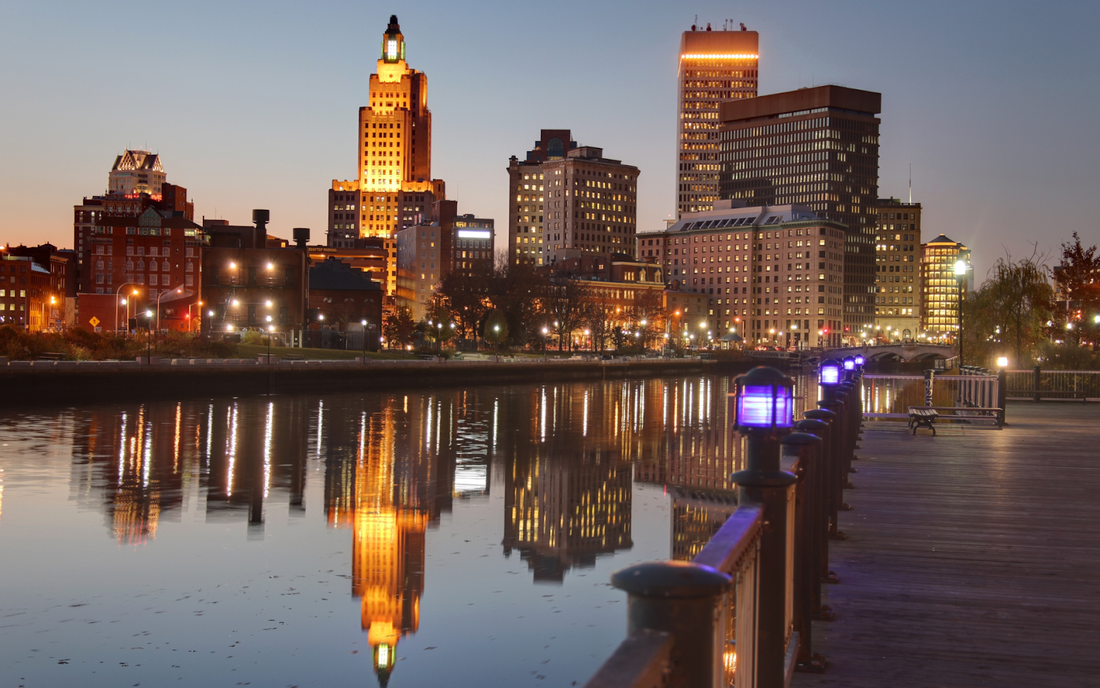 Providence, Rhode Island skyline at dusk reflected in the river with a waterfront boardwalk and purple-lit railing lamps