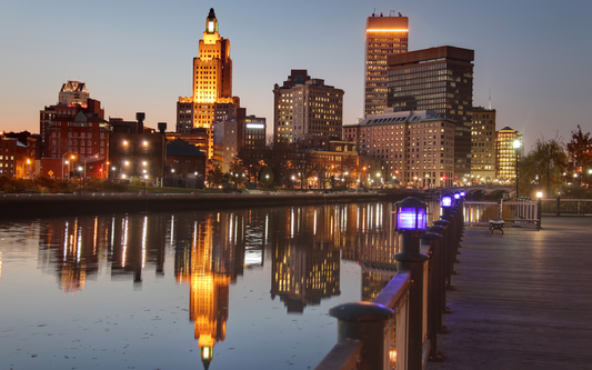 Providence, Rhode Island skyline at dusk reflected in the river with a waterfront boardwalk and purple-lit railing lamps