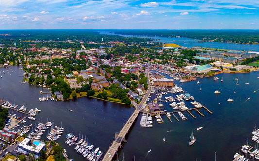 Aerial view of a Maryland waterfront showing marinas, boats, a bridge, and downtown buildings under a blue sky