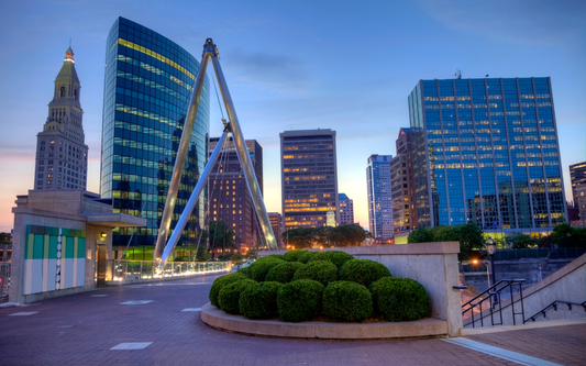 Downtown Connecticut skyline at dusk with lit office towers, a triangular pedestrian bridge, and landscaped shrubs in the foreground