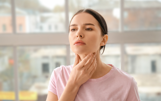 Young woman touching her throat and looking concerned in a bright room, illustrating thyroid discomfort