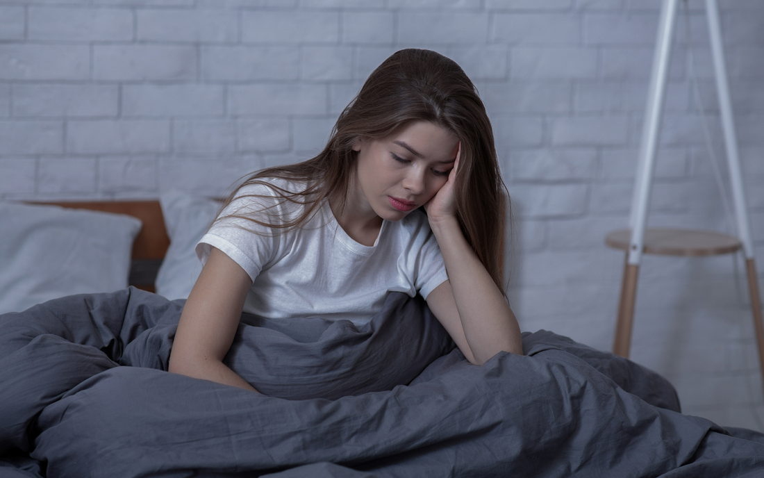 Young woman sitting up in bed holding her head, looking tired and distressed in a dim bedroom