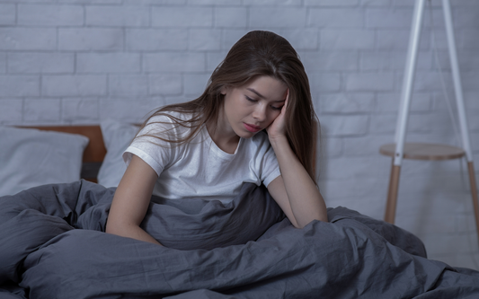 Young woman sitting up in bed holding her head, looking tired and distressed in a dim bedroom