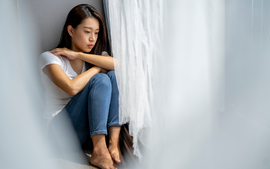 Young woman sitting on a windowsill hugging her knees, looking thoughtful and sad with soft daylight through sheer curtains