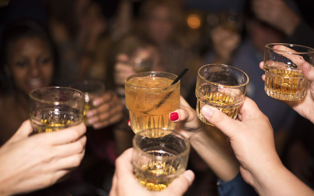 Close-up of several hands clinking whiskey glasses as people toast in a crowded dimly lit bar
