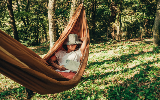 Person napping in a brown hammock in a sunlit forest, bucket hat covering their face and holding a book