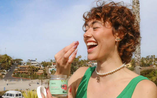 Woman laughing while taking a capsule from a jar of Soul Big Brain focus capsules on a sunny beach